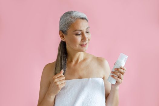 Beautiful grey haired Asian lady wrapped with white towel holds bottle of body care product on pink background in studio. Mature beauty lifestyle