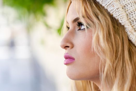 Close-up profile of blonde young woman with curly hair. Girl wearing sweater and wool cap in urban background. Female with beautiful lips painted in pink