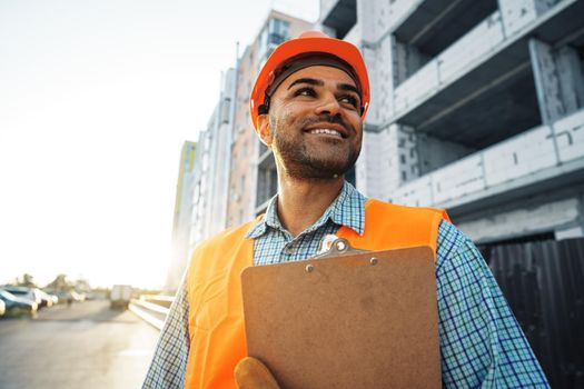 Young man engineer in workwear standing in construction site with clipboard, close up portrait