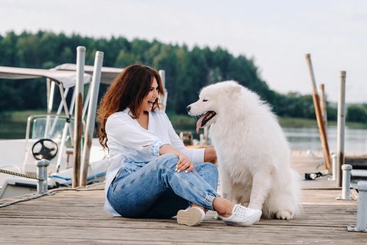 a happy woman with a big white dog sits on a pier by the sea at sunset.