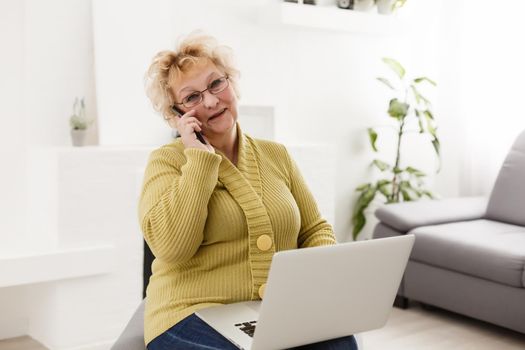 Attractive elderly woman sitting on couch at home, looking at camera, makes video call, communication distantly, modern wireless technologies concept