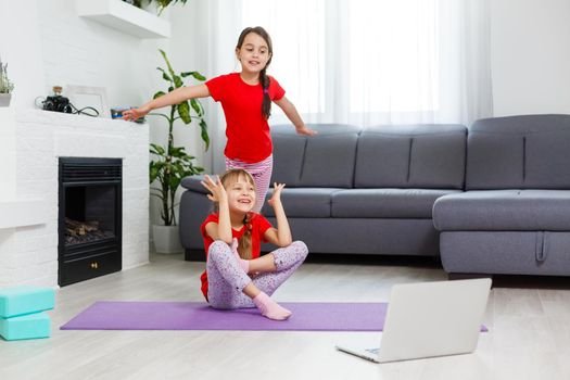 two little girls practicing yoga, stretching, fitness by video on notebook. Distant online education training, aerobic at home. Healthy lifestyle, coronavirus, stay home. Kids sport home quarantine.