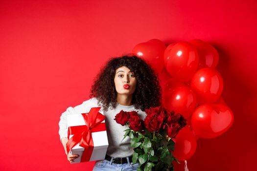 Holidays and celebration. Pretty woman celebrating birthday blowing air kiss, receive gifts and flowers on anniversary, standing near party balloons, red background.
