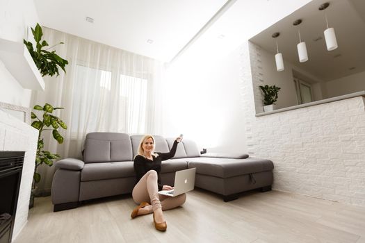 Young woman switching on air conditioner while sitting on sofa near white wall