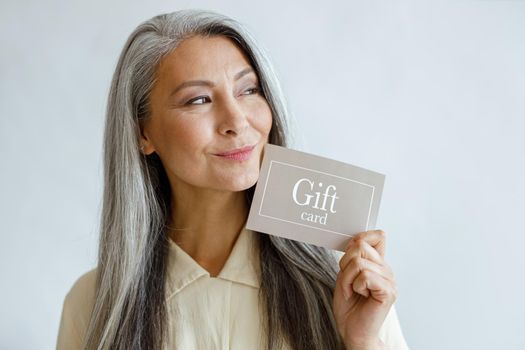 Positive silver haired Asian woman customer holds gift card standing on light grey background in studio. Shopping certificate
