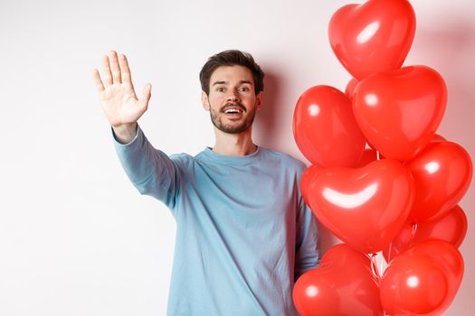 Handsome young caucasian man standing with romantic heart balloon and waving hand at lover, waiting for his date on Valentines day with cute surprise, standing over white background.