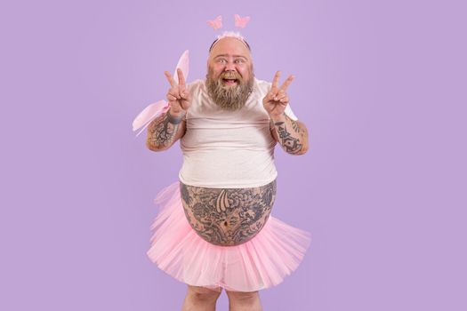 Excited middle aged man with overweight in carnival fairy suit with pink wings shows peace gestures on purple background in studio