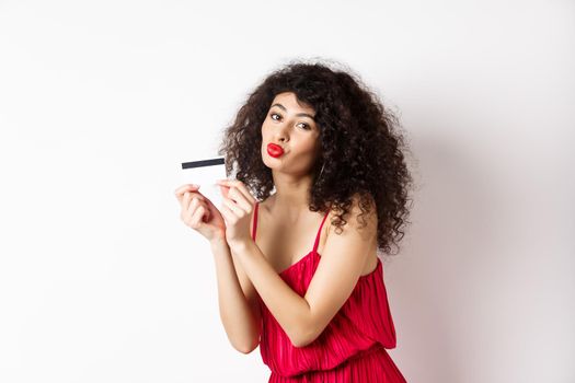 Shopping. Beautiful lady with curly hair, pucker lips, kissing plastic credit card, standing in red dress against white background.