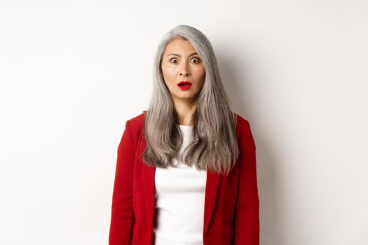 Business concept. Shocked senior female employer staring at camera speechless, standing in red blazer and makeup over white background.