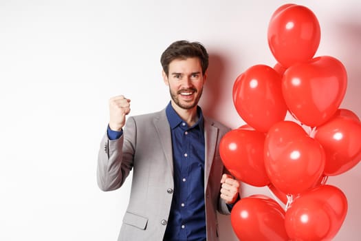 Lovers day. Cheerful young man in suit, feeling confident about Valentines date, saying yes and smiling, standing near hearts balloon, white background.