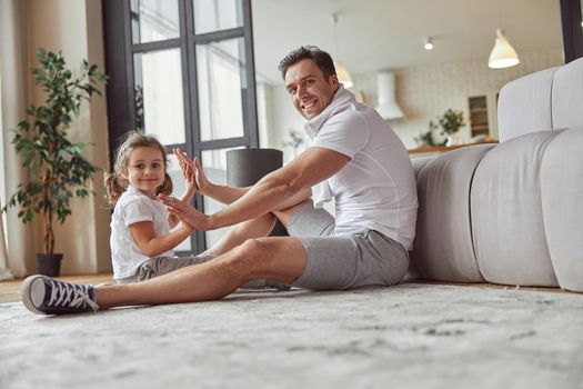 Low angle full length portrait of smiling girl with father sitting on floor in living room and doing patty-cake