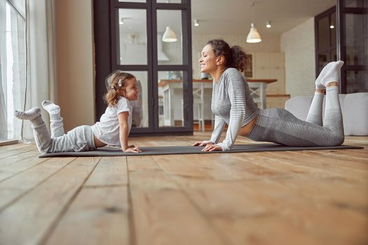 Low angle of cheerful woman doing exercises together with cute girl on floor for improving flexibility