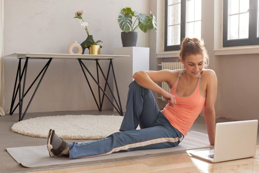 Smiling athletic female in sportswear sitting on mat with laptop during online workout at home and looking at screen