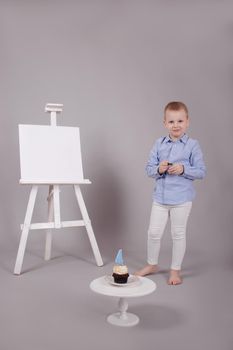 preschool boy in white pants and blue shirt on grey background near easel with mockup and cupcake with candle, shaped in number four 4. happy birthday. celebration. High quality photo