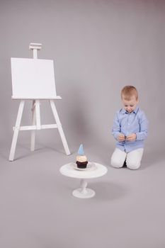 preschool boy in white pants and blue shirt on grey background near easel with mockup and cupcake with candle, shaped in number four 4. happy birthday. celebration. High quality photo