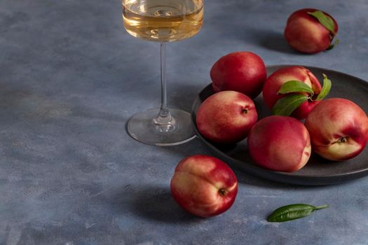 Several ripe peaches or nectarines on a black ceramic plate and a glass of white wine sit on a blue plaster-textured surface. Selective focus. Copy space.