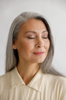 Calm middle aged woman with closed eyes and everyday makeup stands on light background in studio. Mature beauty lifestyle