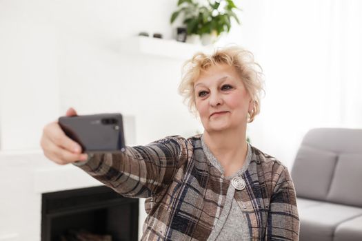 Attractive elderly woman sitting on couch at home, looking at camera, makes video call, communication distantly, modern wireless technologies concept