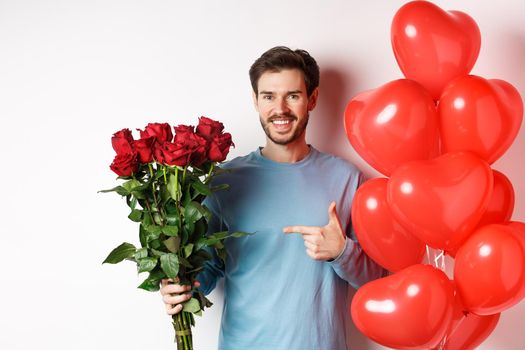 Valentines day romance. Happy young man with flowers and heart balloons going on date with lover. Smiling guy pointing at bouquet of red roses, standing over white background.