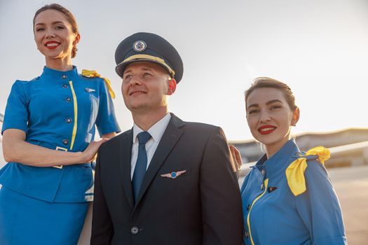 Portrait of male pilot wearing hat posing together with two air hostesses in blue uniform outdoors. Aircraft, occupation concept