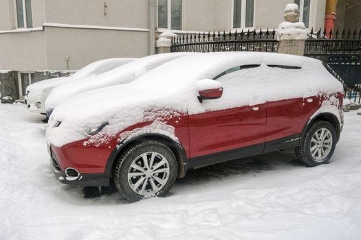 Modern red automobile covered with white snow parked near house on street in wintertime