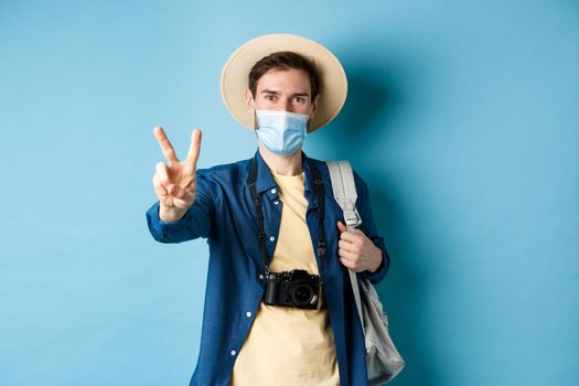 Covid-19, pandemic and travel concept. Positive guy going on vacation, wearing summer hat and medical mask, showing peace sign, standing with backpack and camera on blue background.