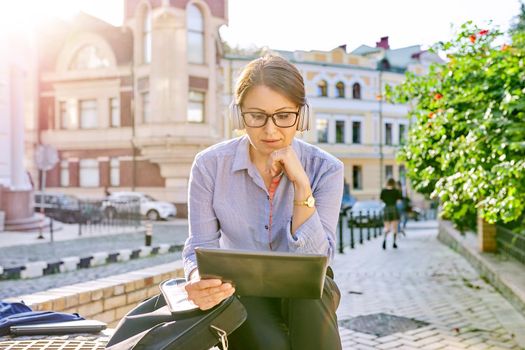 Serious mature business woman in glasses headphones with digital tablet listening attentively and looking at tablet screen, confident female sitting on city street in sunset light