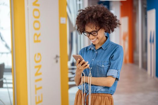 Smiling young woman reading message on her mobile phone while standing in office corridor