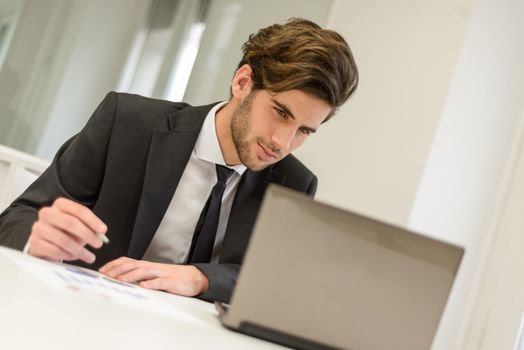 Portrait of a smiling businessman sitting at his laptop and working in a modern office with white furniture