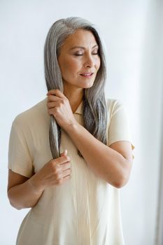 Relaxed middle aged Asian lady in beige blouse strokes lock of silver hair standing on light background in studio. Mature beauty lifestyle