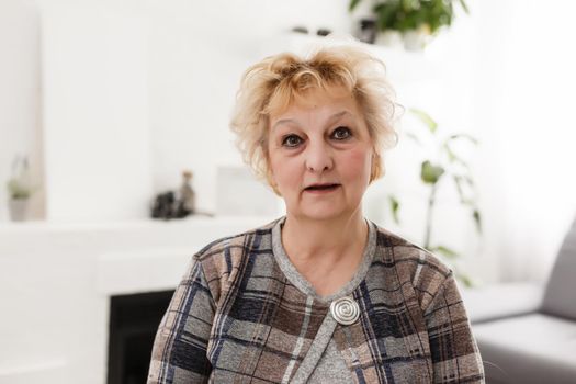 Attractive elderly woman sitting on couch at home, looking at camera, makes video call, communication distantly, modern wireless technologies concept