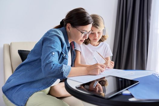 Mom and daughter of school elementary age writing and reading lessons together, parent helping the child, sitting at home on couch at table