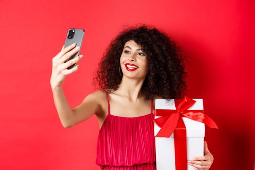 Beautiful girlfriend with curly hair, wearing evening dress, taking selfie with gift from lover, photographing on smartphone and smiling, standing over red background.