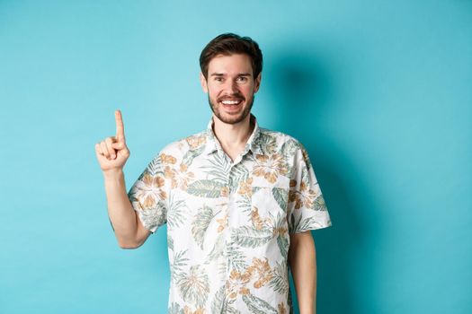 Happy tourist in hawaiian shirt smiling and pointing finger up, showing logo promotion, standing on blue background.