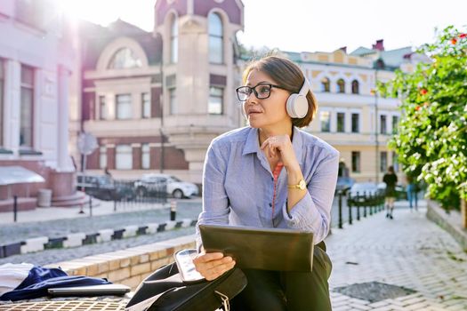 Serious mature business woman in glasses headphones with digital tablet listening attentively and looking at tablet screen, confident female sitting on city street in sunset light