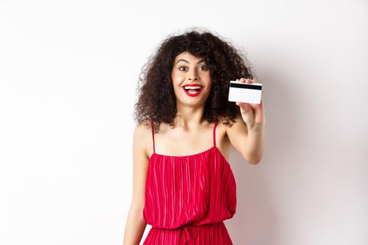 Shopping. Excited stylish lady with makeup, red dress, showing plastic credit card and smiling amazed, standing against white background.