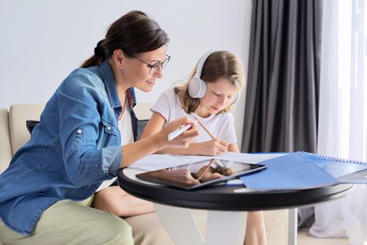 Mom and daughter of school elementary age writing and reading lessons together, parent helping the child, sitting at home on couch at table