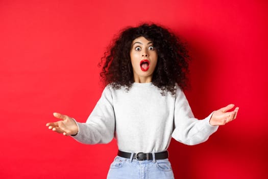 Beautiful young woman standing in awe, drop jaw and looking impressed at camera, spread hands sideways with disbelief, standing on red background.