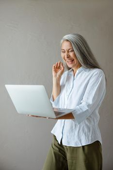 Joyful silver haired mature Asian woman laughs communicating at videocall via modern laptop near grey stone wall in studio