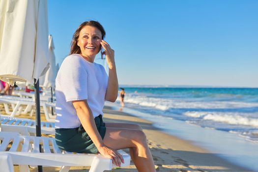Profile view of mature woman talking on phone on sea beach at evening sunset time, copy space on sea natural landscape, horizon, blue sky. Vacation, weekend, relaxation, middle-aged people