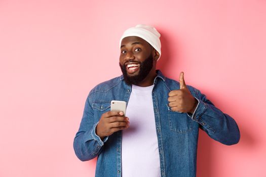 Technology and online shopping concept. Satisfied Black man smiling, using mobile phone and showing thumb-up in approval, making order, standing over pink background.