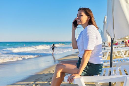 Profile view of mature woman talking on phone on sea beach at evening sunset time, copy space on sea natural landscape, horizon, blue sky. Vacation, weekend, relaxation, middle-aged people