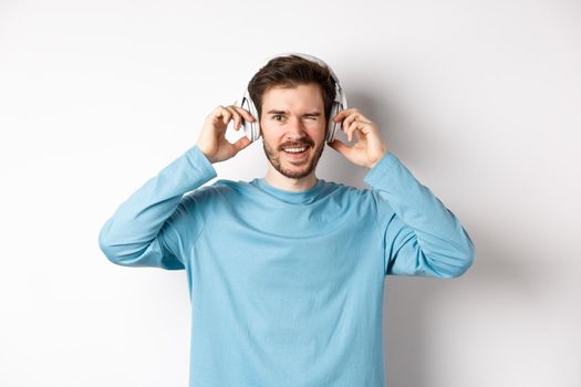 Cheerful guy wink at camera and smiling, listening music in wireless headphones, white background.