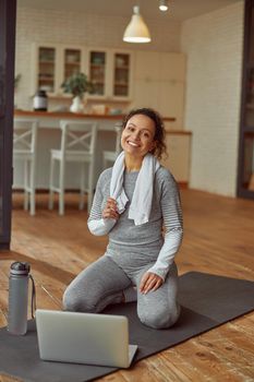 Full length portrait of smiling female with towel sitting on mat during training with notebook at home