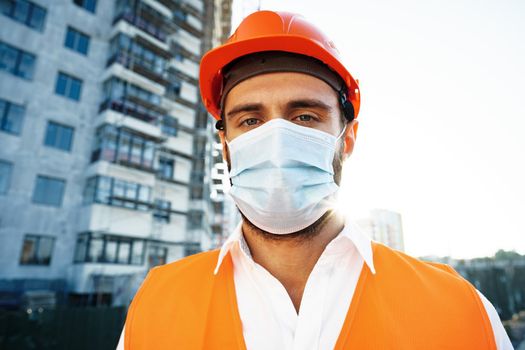 Portrait of man builder in workwear and hardhat wearing medical mask, close up photo