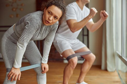 Woman is feeling tired while exercising with husband and doing squats with resistance bands indoors