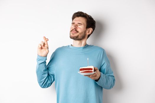 Celebration and holidays concept. Handsome young man celebrating birthday, making wish with bday cake in hand, standing over white background.