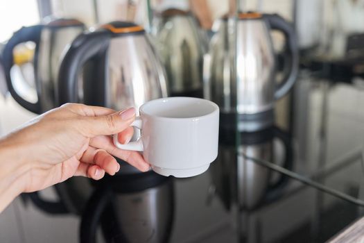 White coffee cup in hand close-up, stainless steel electric kettle in the mirror background