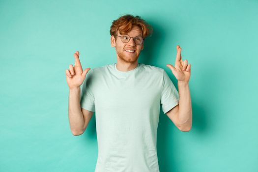 Hopeful redhead man in glasses making a wish, cross fingers and looking right at copy space, dreaming about something or making wish, standing over turquoise background.