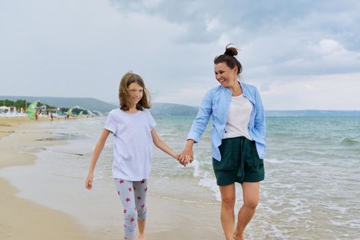 Happy mom and daughter walking on the sea beach holding hands. Family middle-aged mother and preteen child together, vacation, sea weekend, travel, love, happiness and joy